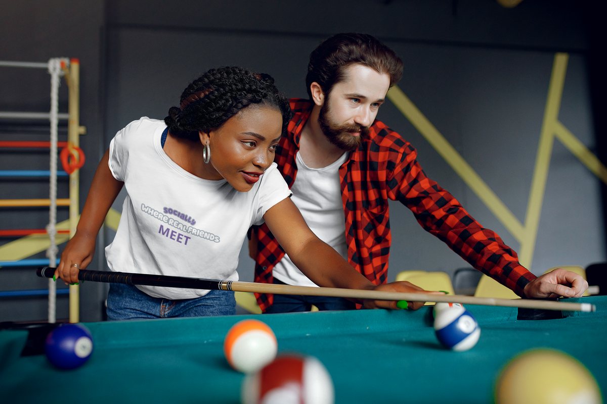 Two friends leaning over a pool table mid-game, one wearing a Socially tee